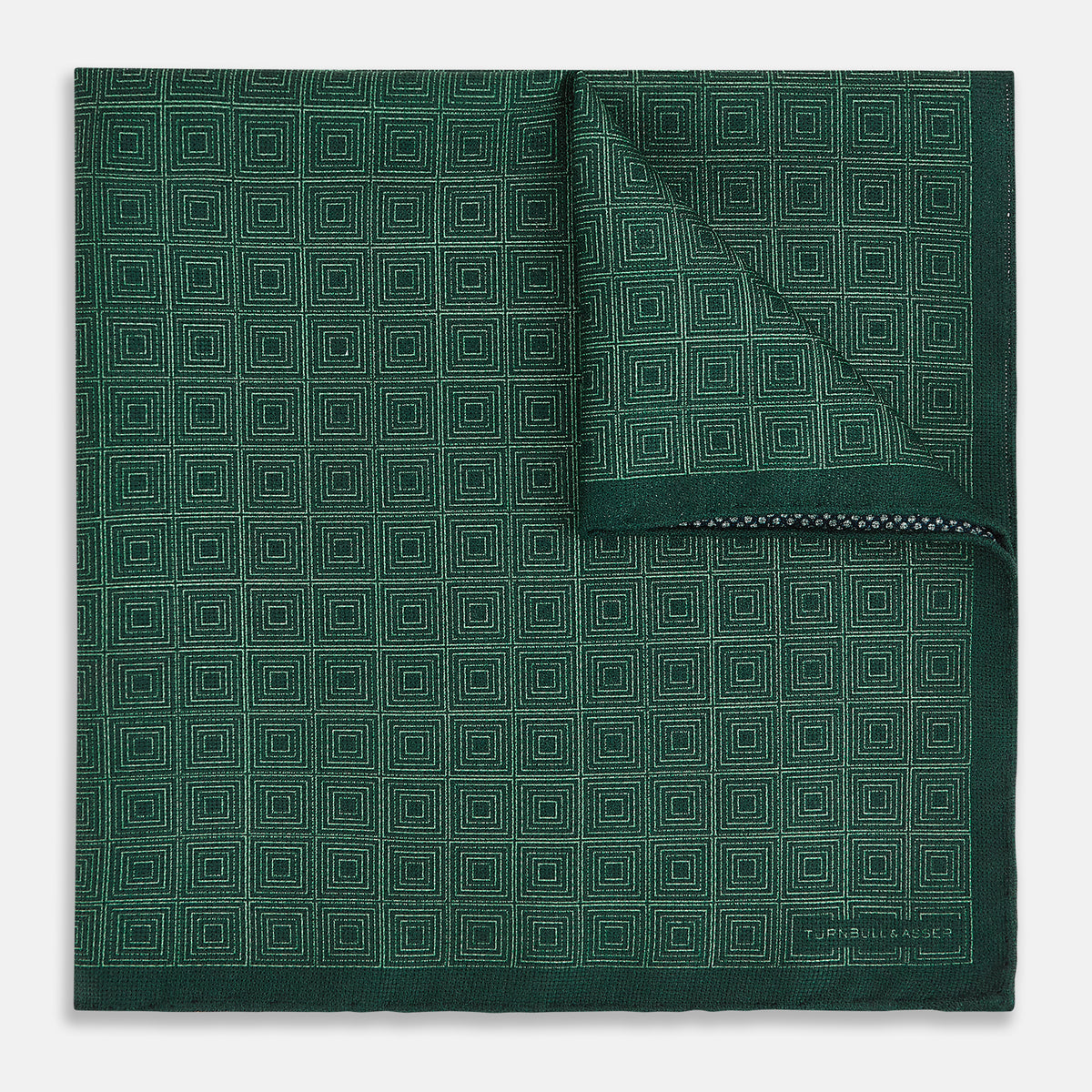View 1 of product The Geometric Green Tile Pocket Square in Silk by Turnbull & Asser UK features a sophisticated tonal green design with a subtle pattern of concentric squares. One corner is folded to highlight the elegant double-sided construction, revealing a contrasting black and white small dot pattern on the reverse.