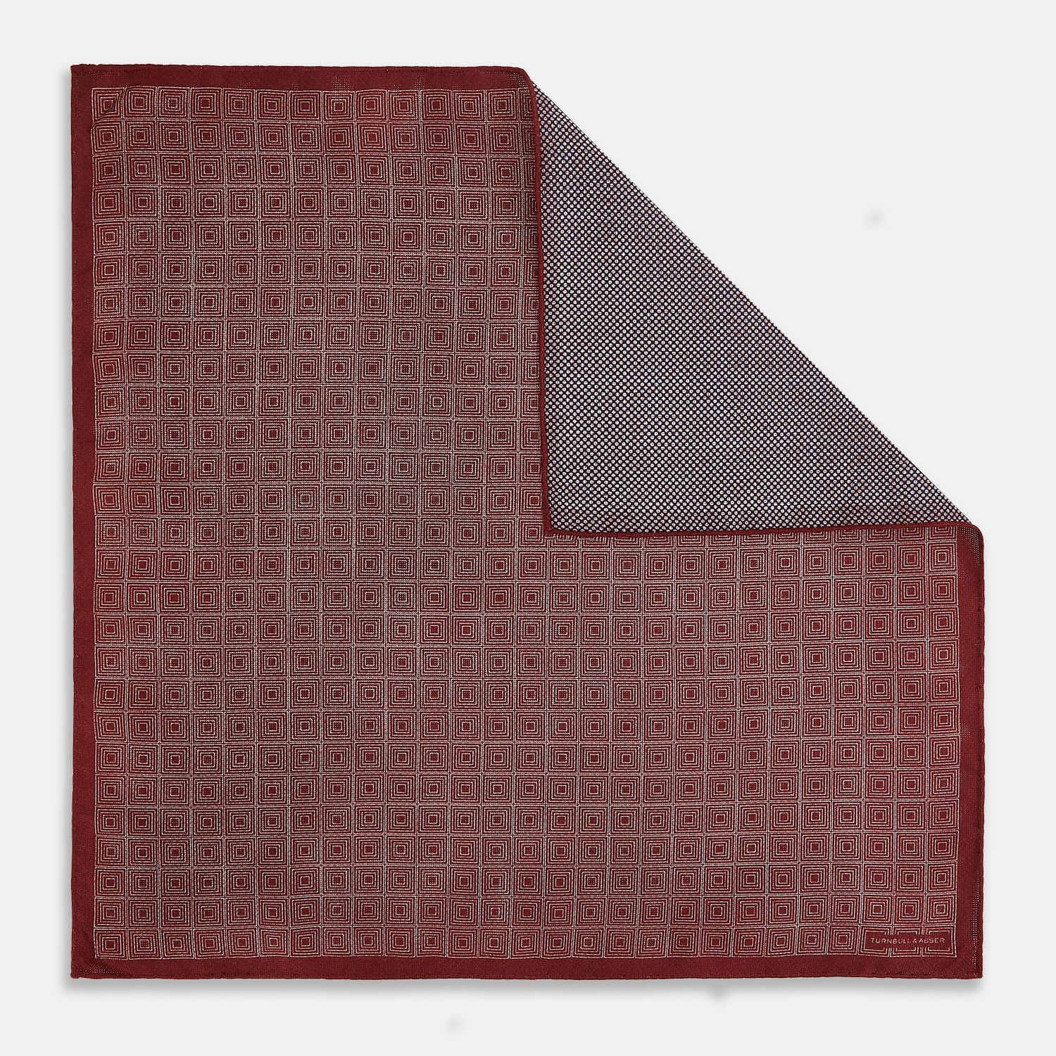 View 1 of product The Turnbull & Asser UK Geometric Burgundy Tile Pocket Square in Silk is displayed partly folded, showcasing its bold geometric grid pattern with concentric burgundy squares on the front and a contrasting black-and-white dotted design on the reverse.