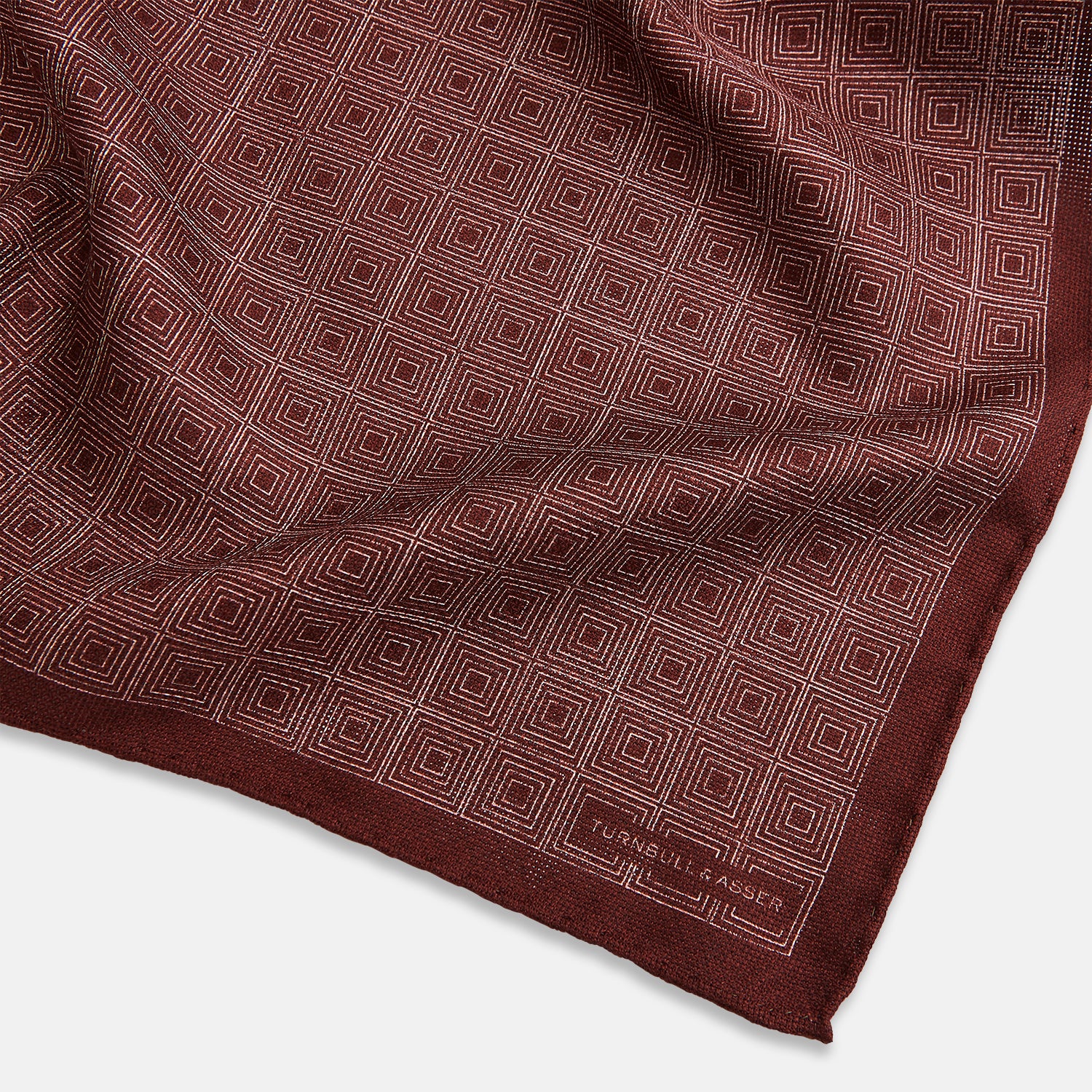 View 3 of product The Turnbull & Asser UK Geometric Burgundy Tile Pocket Square in Silk is displayed partly folded, showcasing its bold geometric grid pattern with concentric burgundy squares on the front and a contrasting black-and-white dotted design on the reverse.