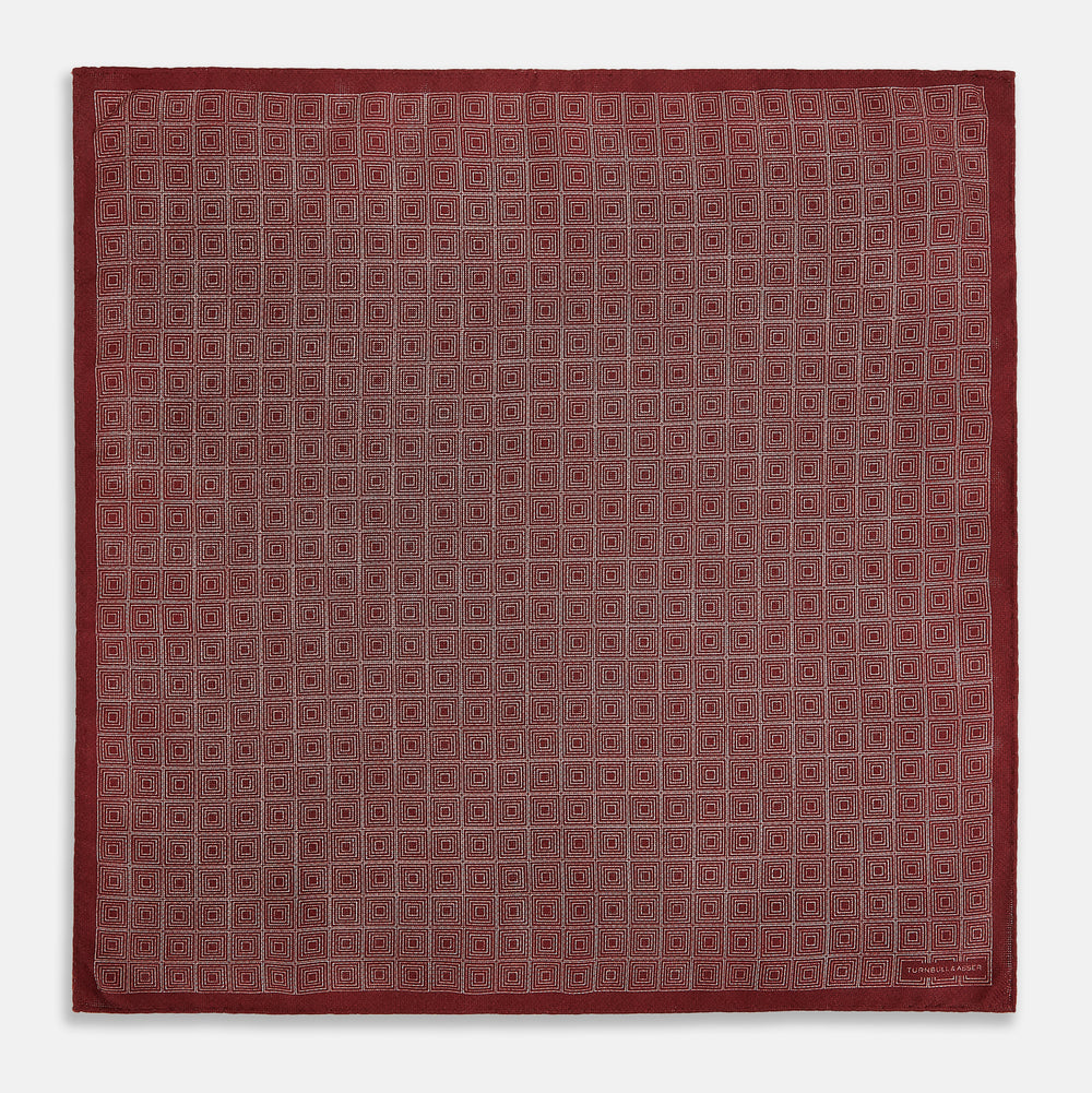The Turnbull & Asser UK Geometric Burgundy Tile Pocket Square in Silk is displayed partly folded, showcasing its bold geometric grid pattern with concentric burgundy squares on the front and a contrasting black-and-white dotted design on the reverse.