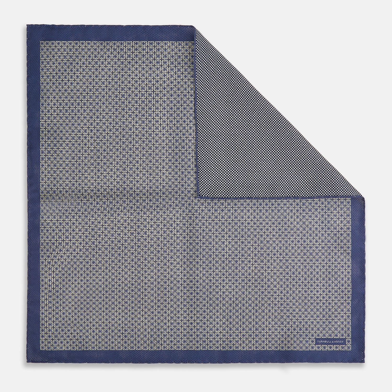 View 1 of product The Geometric Blue Pocket Square in Silk from Turnbull & Asser UK showcases a blue border and double-sided design, with a geometric pattern at the center. A folded corner reveals a refined, tighter dot motif on the reverse side, emphasizing the intricate craftsmanship of this finely woven accessory.