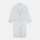 The Turnbull & Asser White Piped Herringbone Cotton Gown features long sleeves, a waist tie, chest pocket, and striking red piping on the collar, cuffs, pocket, and edges—an elegant piece showcased against a white background.