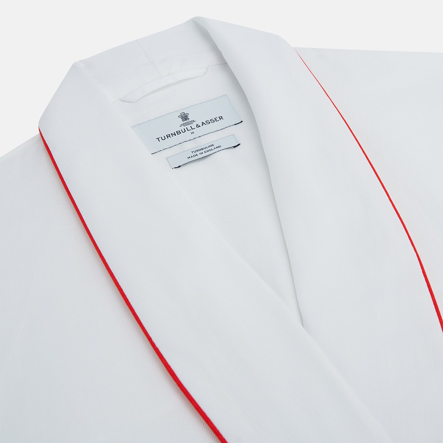 View 2 of product The Turnbull & Asser White Piped Herringbone Cotton Gown features long sleeves, a waist tie, chest pocket, and striking red piping on the collar, cuffs, pocket, and edges—an elegant piece showcased against a white background.