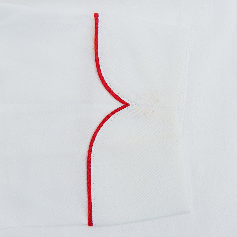 The Turnbull & Asser White Piped Herringbone Cotton Gown features long sleeves, a waist tie, chest pocket, and striking red piping on the collar, cuffs, pocket, and edges—an elegant piece showcased against a white background.