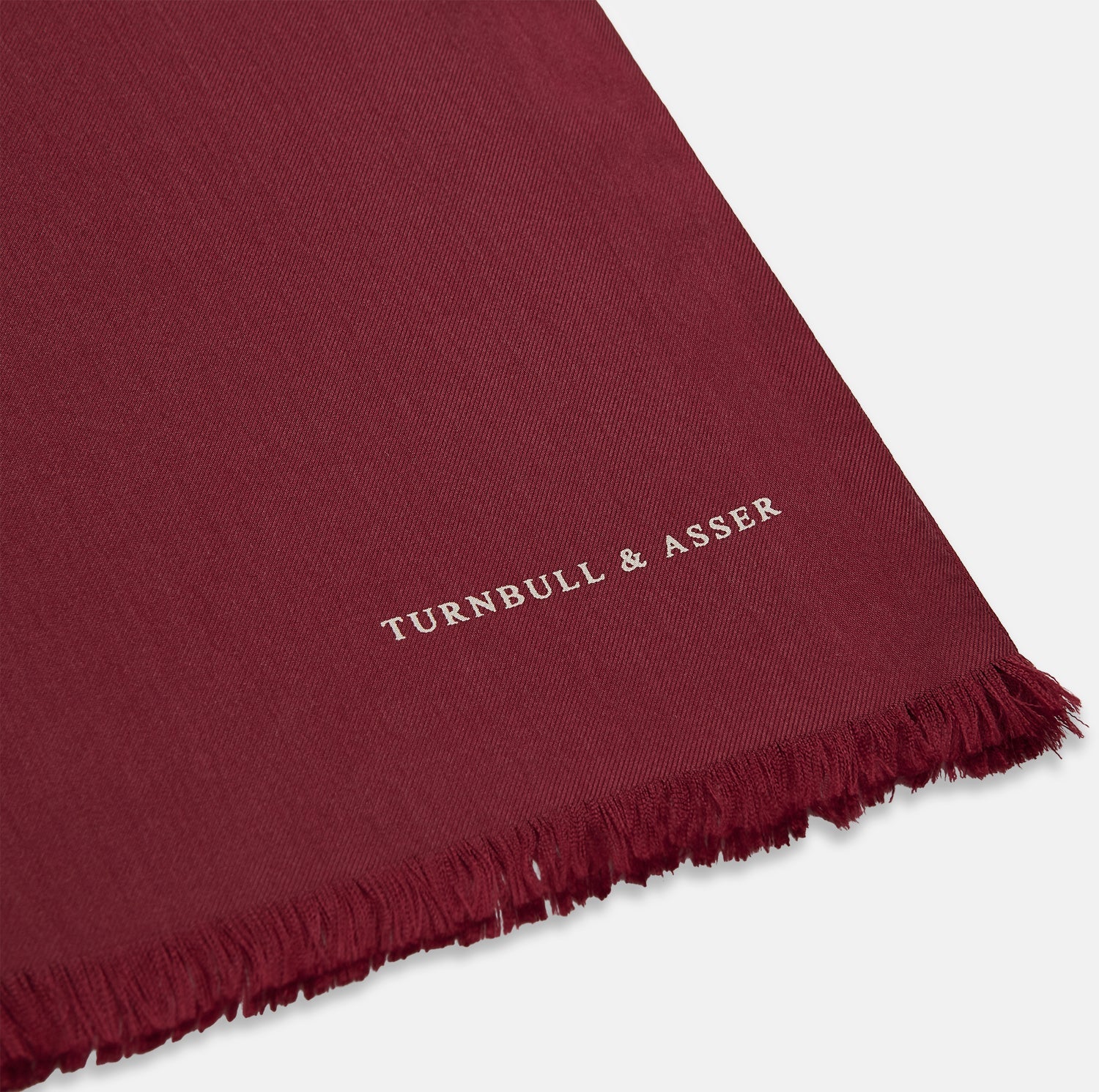 View 2 of product The Plain Wine Silk Scarf by Turnbull & Asser is a long, smooth accessory with subtle fringe and white Turnbull & Asser embroidery on one corner. It comes neatly folded in a deep wine color, perfect for an elegant touch.