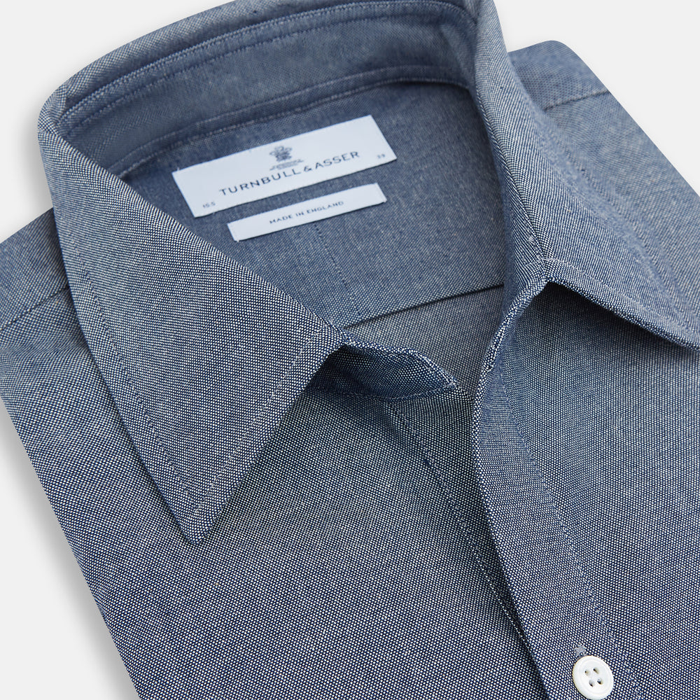The Turnbull & Asser Steel Blue Piccadilly Shirt features long sleeves, a button-up front, two chest pockets, and white buttons, shown against a plain white background.