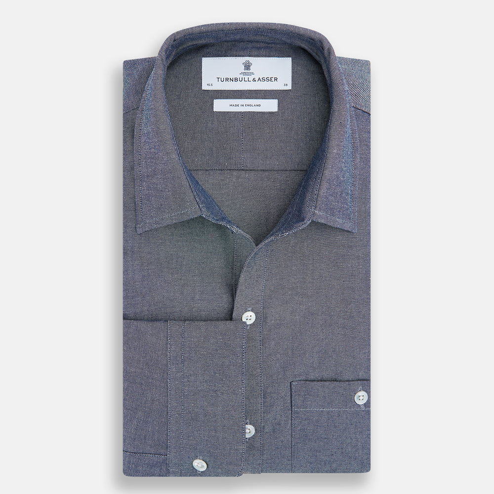 The Turnbull & Asser Steel Blue Piccadilly Shirt features long sleeves, a button-up front, two chest pockets, and white buttons, shown against a plain white background.