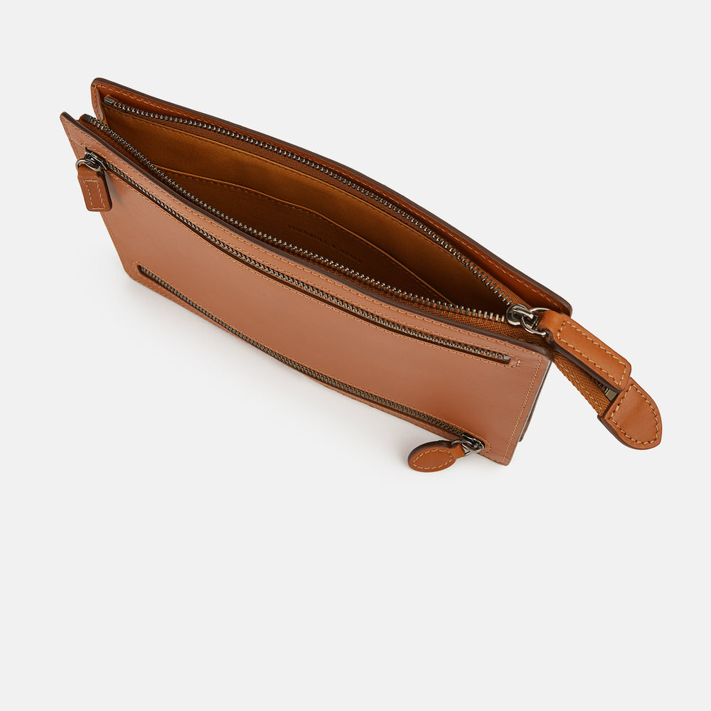 The Cognac Leather Zip Envelope Pouch by Turnbull & Asser UK features two horizontal zippered pockets, stitched edges, and leather pulls—offering a stylish way to organize essentials when you travel.