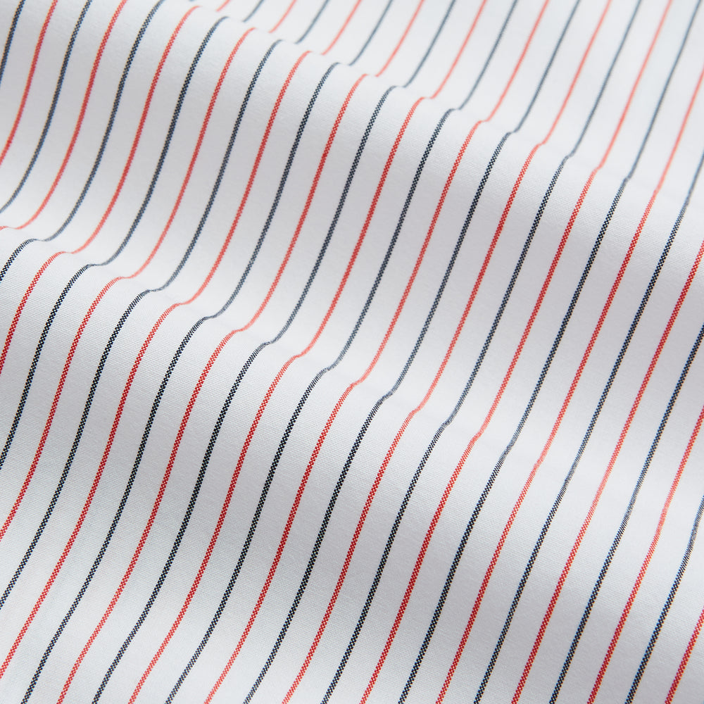 The Turnbull & Asser Red and Navy Pinstripe Mayfair Shirt, an organic cotton long-sleeve button-up with a collar, features thin vertical red and navy pinstripes on white, shown laid flat on a white surface.
