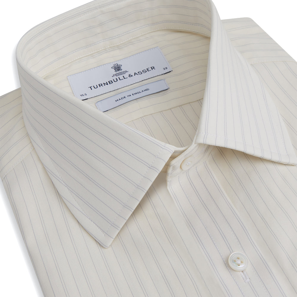 The Turnbull & Asser Pale Blue Fine Track Stripe Mayfair Shirt is a long-sleeve, cream-colored organic cotton shirt with subtle pale blue stripes, a T&A collar, and buttoned cuffs, shown on a plain white background.