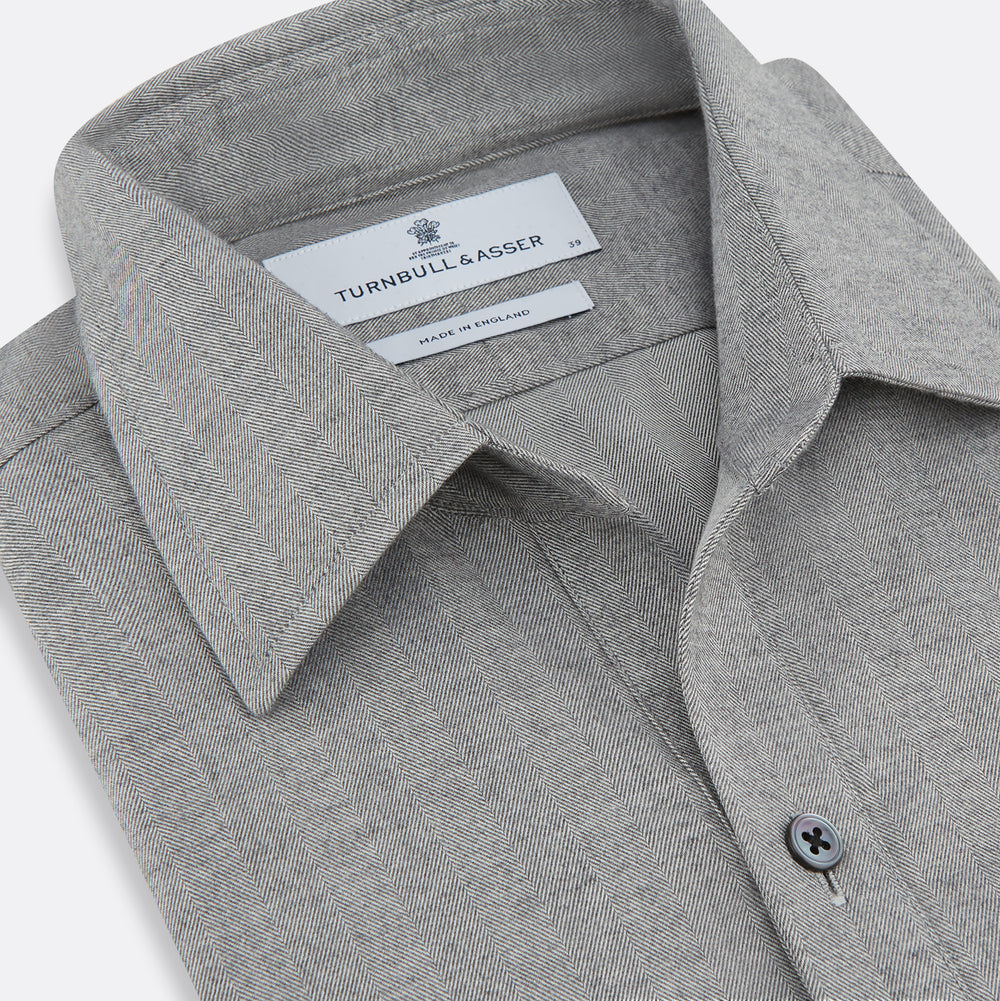 The Turnbull & Asser Grey Herringbone Chelsea Shirt features long sleeves, a chest pocket, classic collar, and buttoned cuffs, adding refined style to smart-casual outfits—shown here against a plain white background.