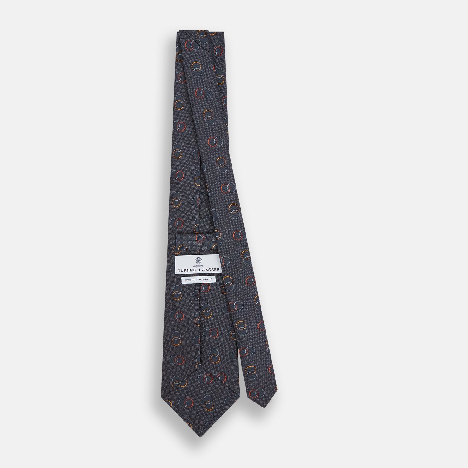 View 4 of product The Navy Links Silk Tie by Turnbull & Asser, featuring a pattern of overlapping red, yellow, and blue rings, is rolled up and displayed against a plain white background.
