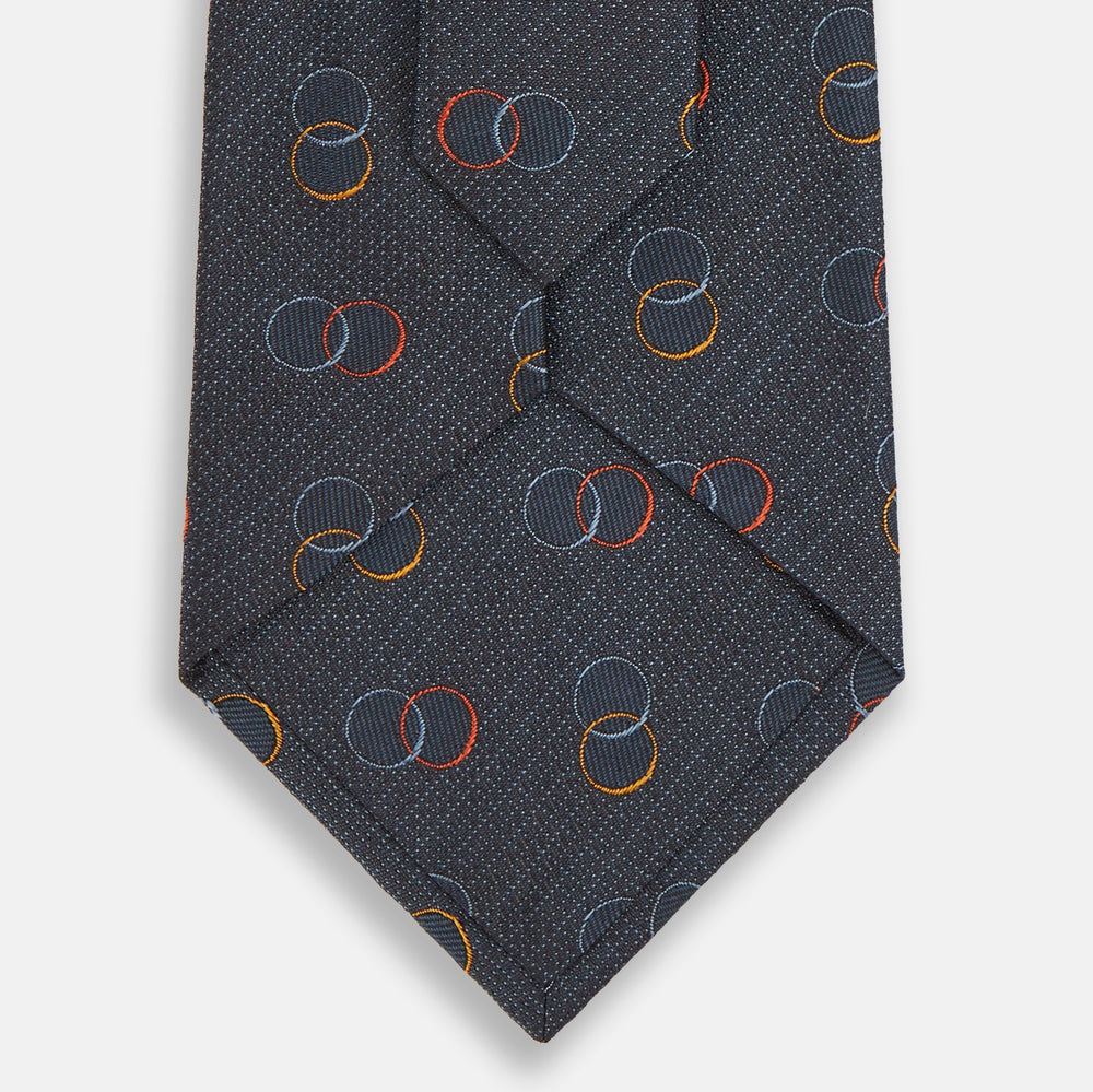 The Navy Links Silk Tie by Turnbull & Asser, featuring a pattern of overlapping red, yellow, and blue rings, is rolled up and displayed against a plain white background.