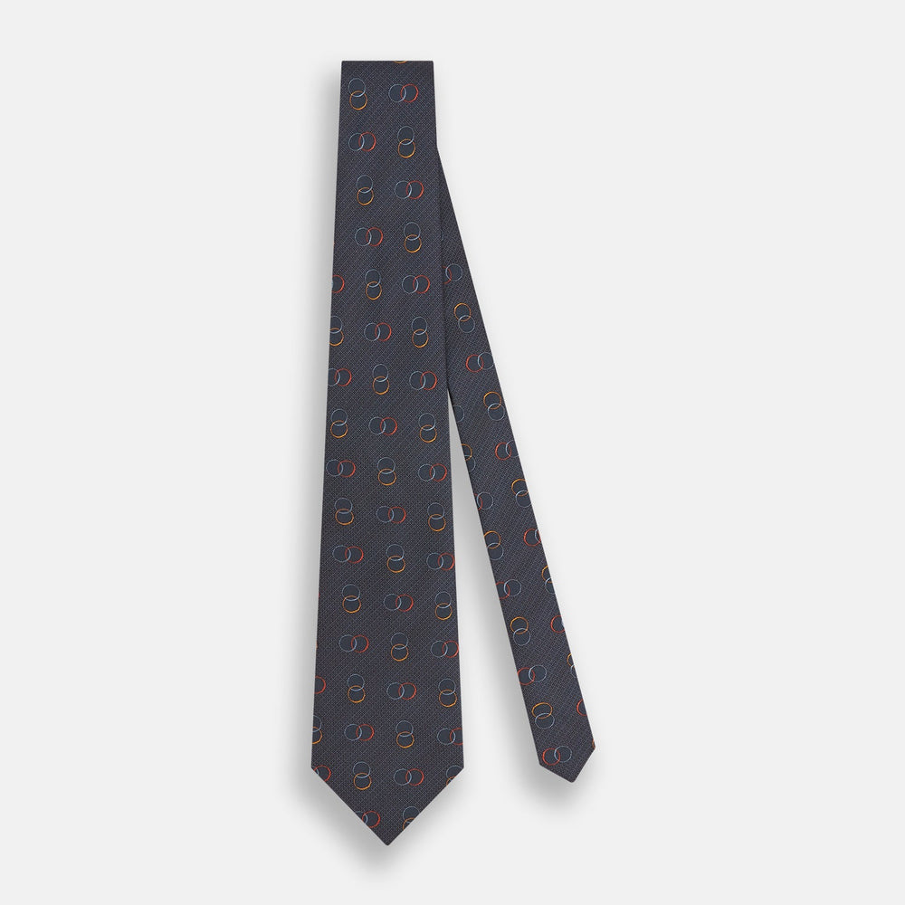 The Navy Links Silk Tie by Turnbull & Asser, featuring a pattern of overlapping red, yellow, and blue rings, is rolled up and displayed against a plain white background.