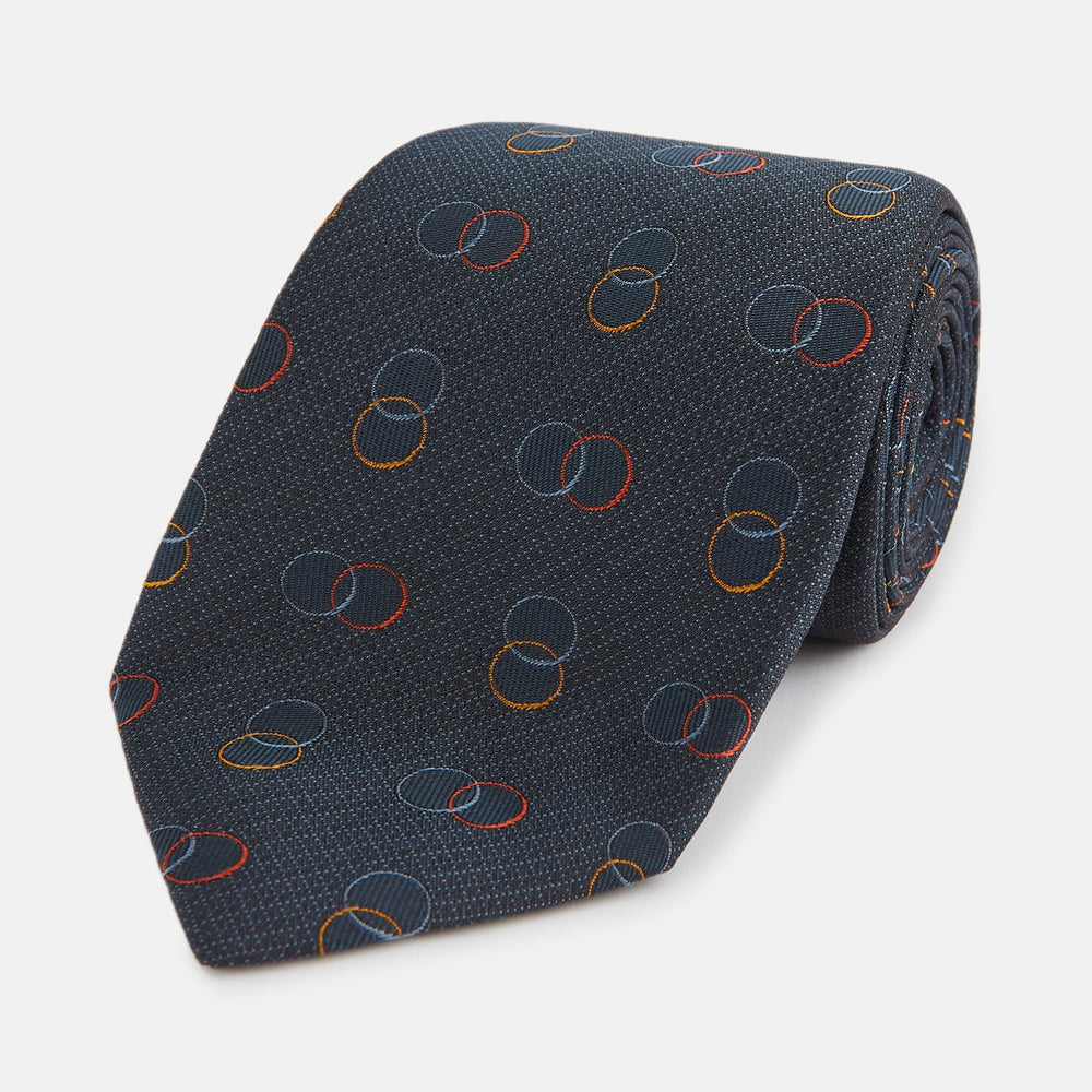 The Navy Links Silk Tie by Turnbull & Asser, featuring a pattern of overlapping red, yellow, and blue rings, is rolled up and displayed against a plain white background.