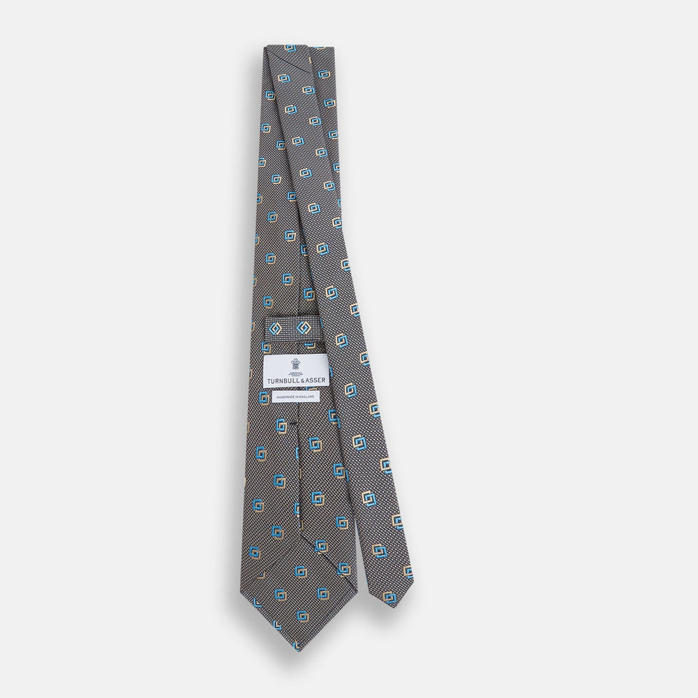 The Turnbull & Asser Blue and Gold Silk Tie, featuring a refined repeating geometric pattern, exudes British elegance and is displayed rolled up on a plain white background.