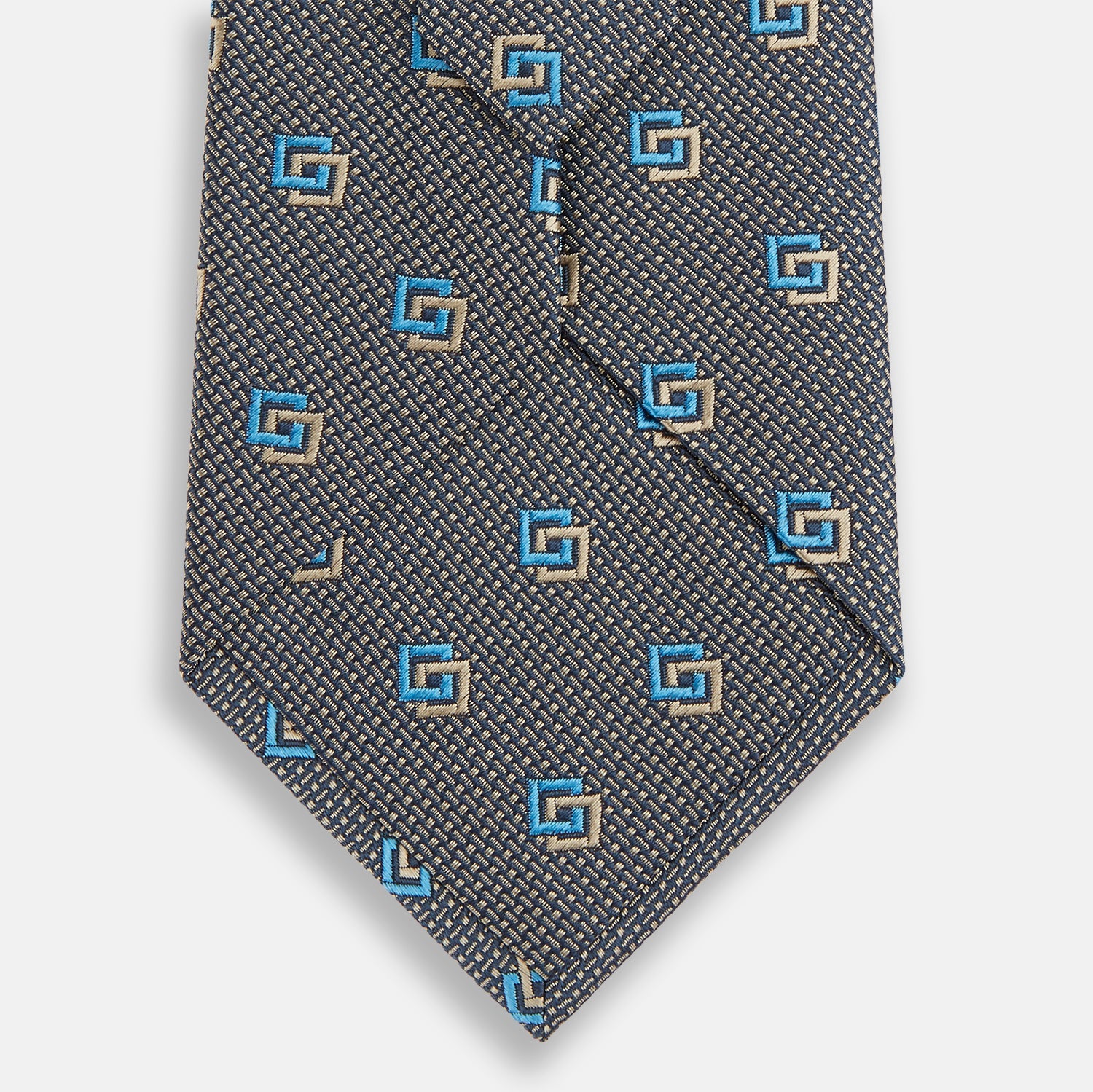 View 3 of product The Turnbull & Asser Blue and Gold Silk Tie, featuring a refined repeating geometric pattern, exudes British elegance and is displayed rolled up on a plain white background.