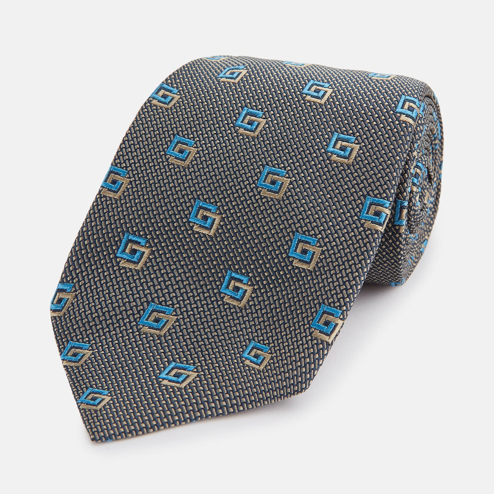 The Turnbull & Asser Blue and Gold Silk Tie, featuring a refined repeating geometric pattern, exudes British elegance and is displayed rolled up on a plain white background.