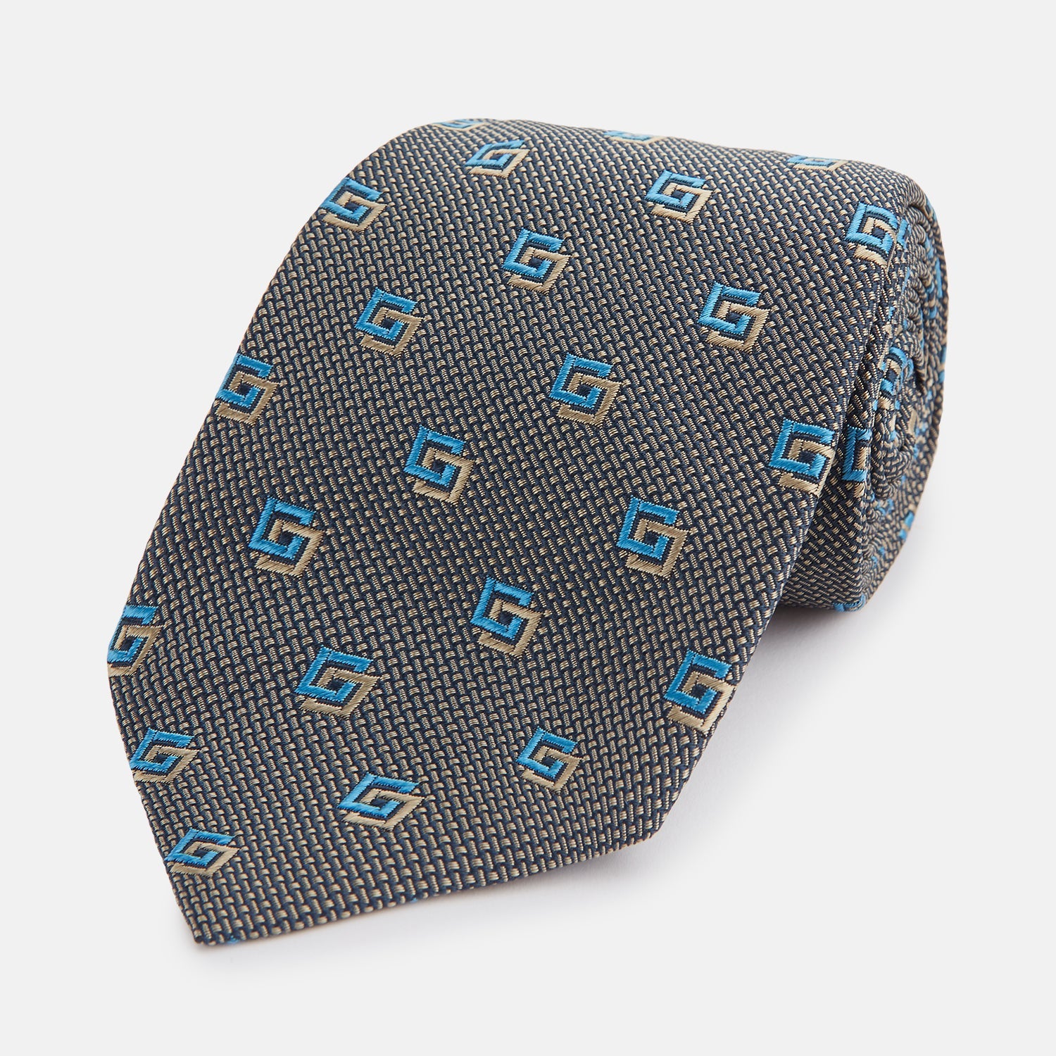 View 1 of product The Turnbull & Asser Blue and Gold Silk Tie, featuring a refined repeating geometric pattern, exudes British elegance and is displayed rolled up on a plain white background.