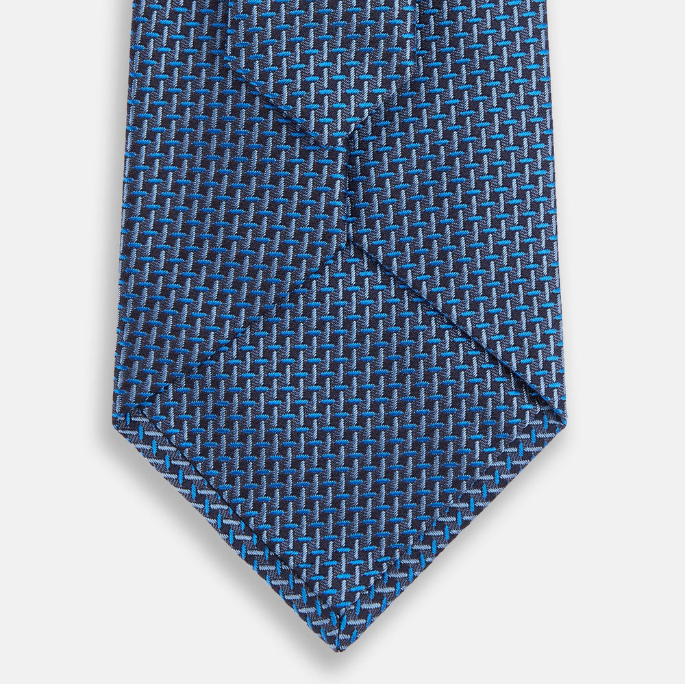 The Turnbull & Asser Blue Geometric Silk Tie features a textured woven pattern with lighter blue accents, shown neatly rolled on a plain white background.