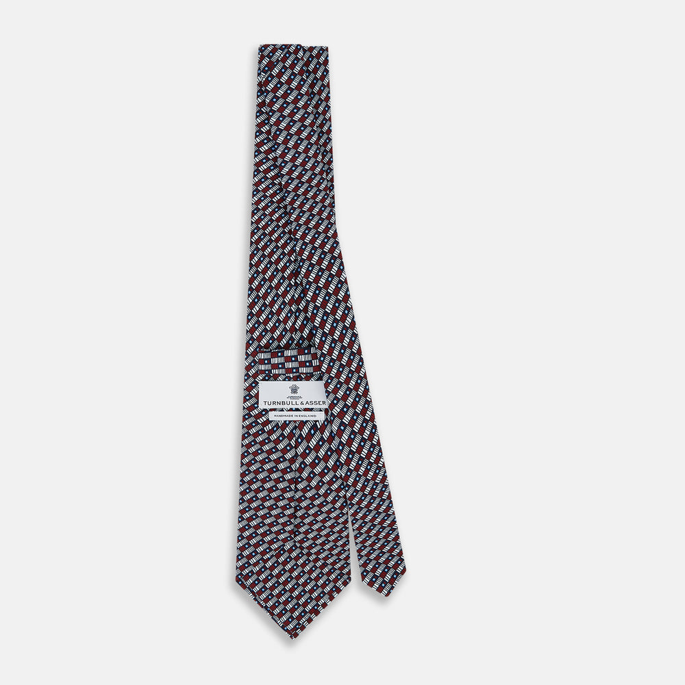 The Turnbull & Asser UK TIES 9.5CM GEOMETRIC RECTANGLES RED is an English silk tie with small red, navy blue, and white rectangles and dots on a white background.