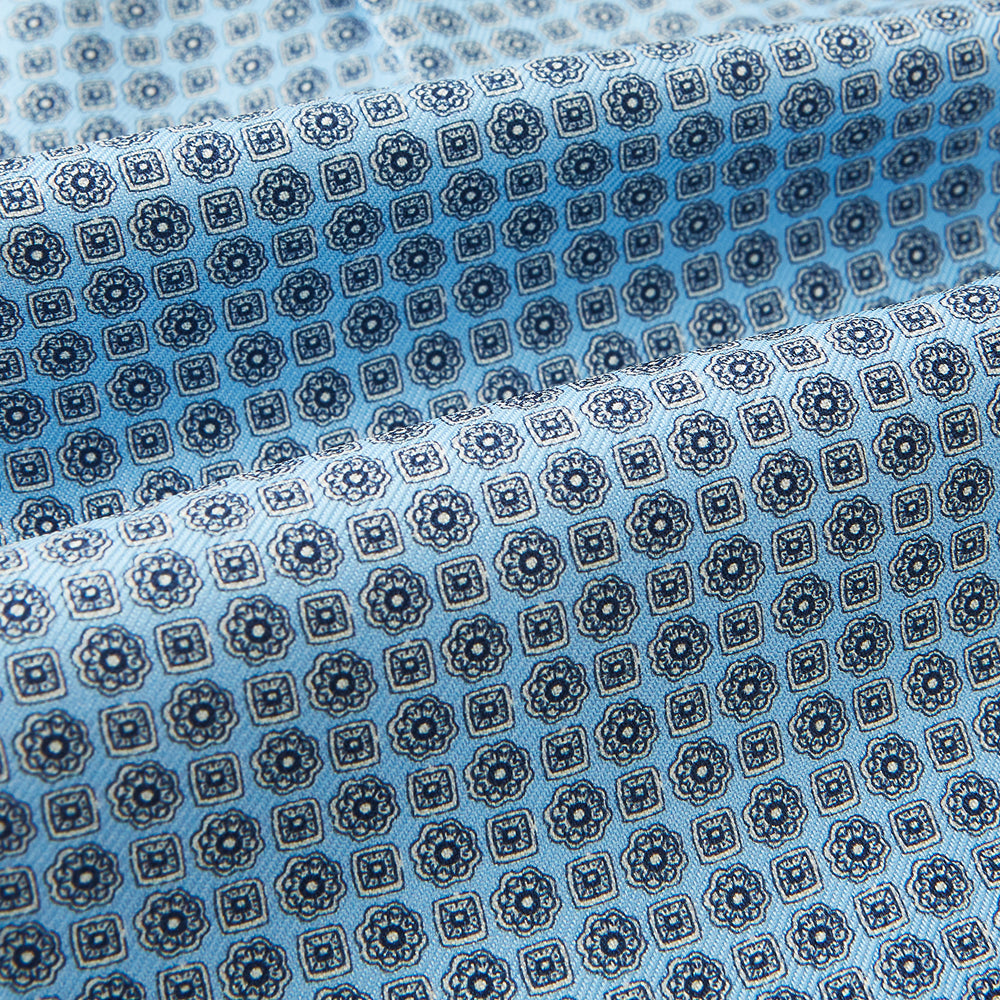 Close-up of Turnbull & Asser UK's Pale Blue Floral Trellis Silk Cravat, featuring light blue silk with a repeating dark blue geometric floral trellis pattern. The fabric appears slightly folded, highlighting its textured, decorative look.