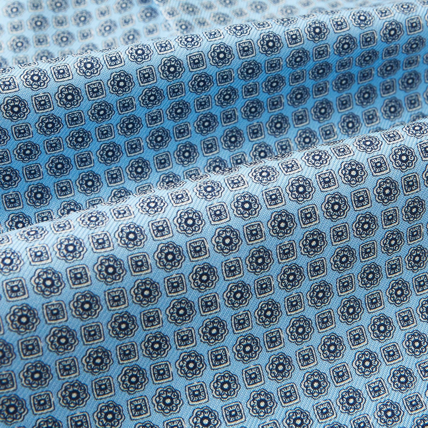 View 4 of product Close-up of Turnbull & Asser UK's Pale Blue Floral Trellis Silk Cravat, featuring light blue silk with a repeating dark blue geometric floral trellis pattern. The fabric appears slightly folded, highlighting its textured, decorative look.