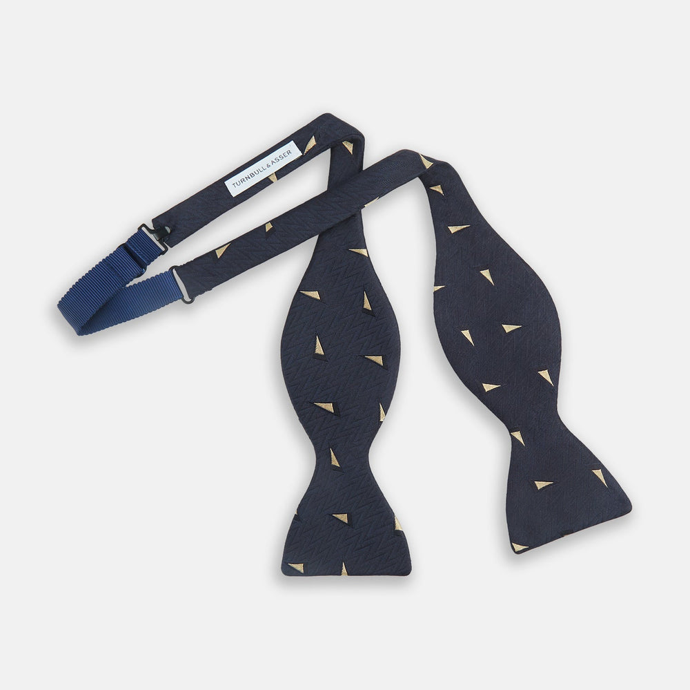 The Navy Triangle Silk Bow Tie by Turnbull & Asser features British elegance with a subtle herringbone pattern and scattered small gold triangle designs.