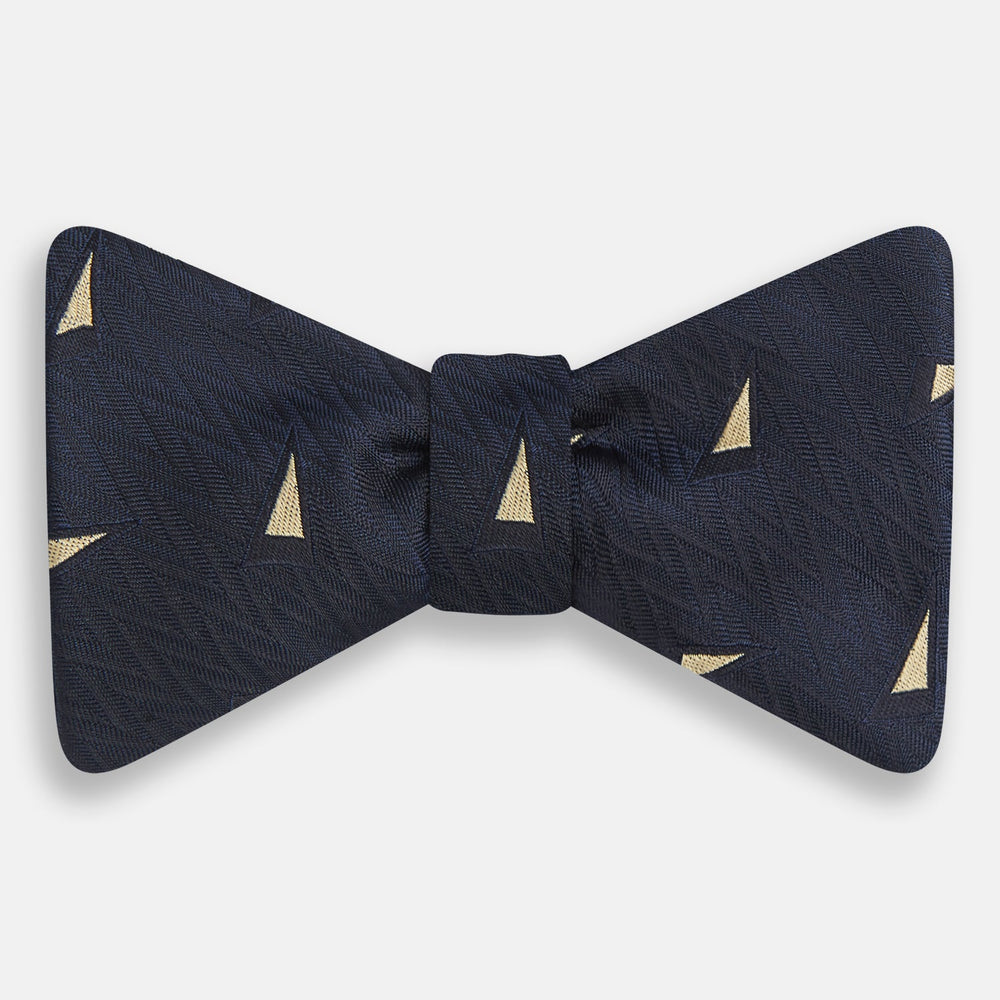 The Navy Triangle Silk Bow Tie by Turnbull & Asser features British elegance with a subtle herringbone pattern and scattered small gold triangle designs.
