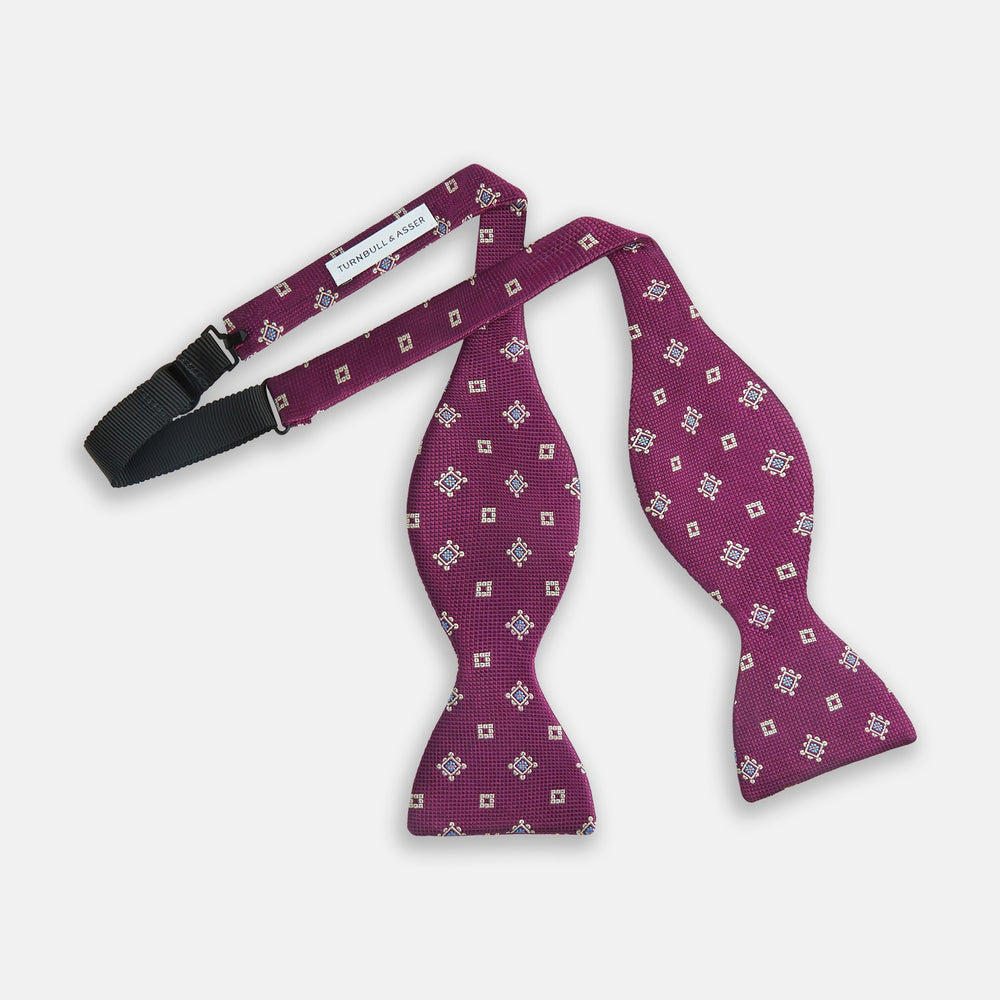 The Magenta Motif Silk Bow Tie by Turnbull & Asser features a textured magenta background with small, square blue, white, and gold geometric designs—an elegant choice for formal occasions.