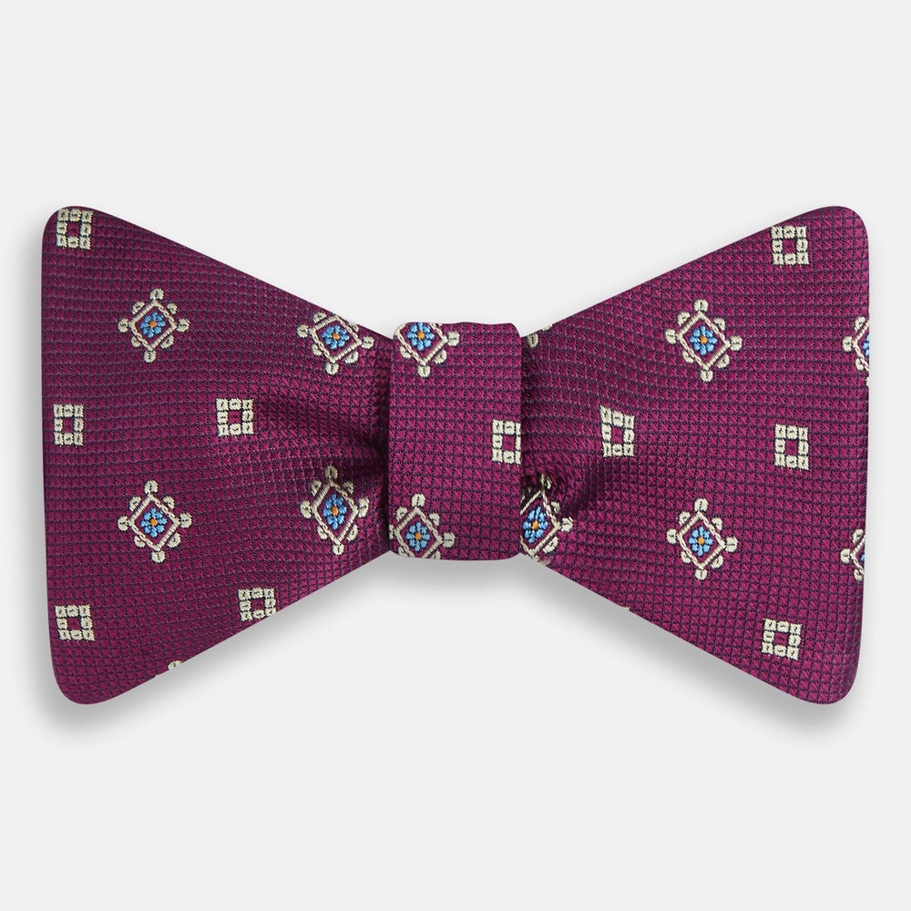 The Magenta Motif Silk Bow Tie by Turnbull & Asser features a textured magenta background with small, square blue, white, and gold geometric designs—an elegant choice for formal occasions.
