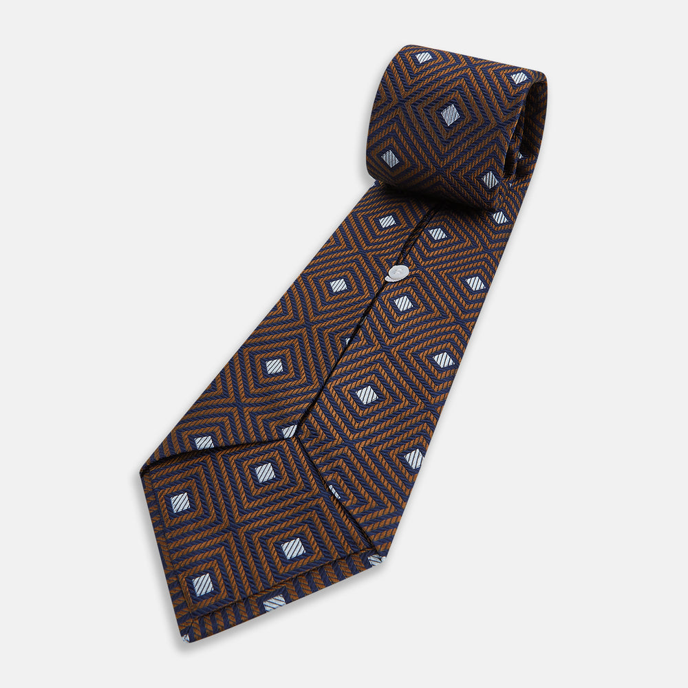 The Tomorrow Never Dies Seven-Fold Silk Tie by Turnbull & Asser UK features a geometric pattern of navy and brown squares with light blue diamond accents, rolled up and displayed on a plain white background.