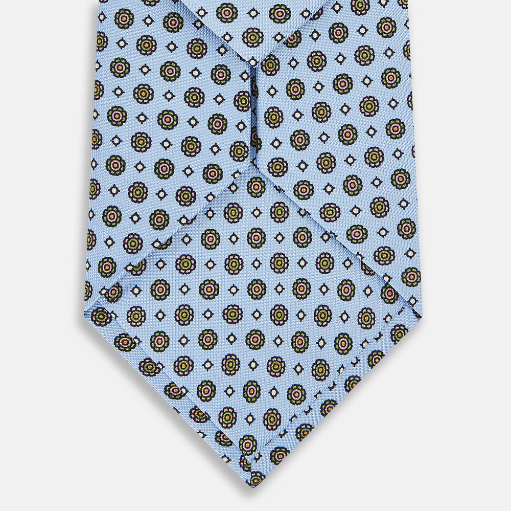 The Turnbull & Asser UK Great Gatsby Seven-Fold Silk Sky Tie, as seen on Jay Gatsby, is a light blue silk tie with a repeating floral medallion pattern in black, yellow, and red circles, shown rolled on a white background.