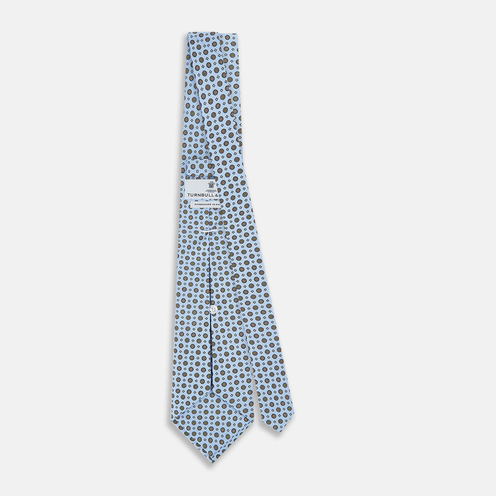 The Turnbull & Asser UK Great Gatsby Seven-Fold Silk Sky Tie, as seen on Jay Gatsby, is a light blue silk tie with a repeating floral medallion pattern in black, yellow, and red circles, shown rolled on a white background.