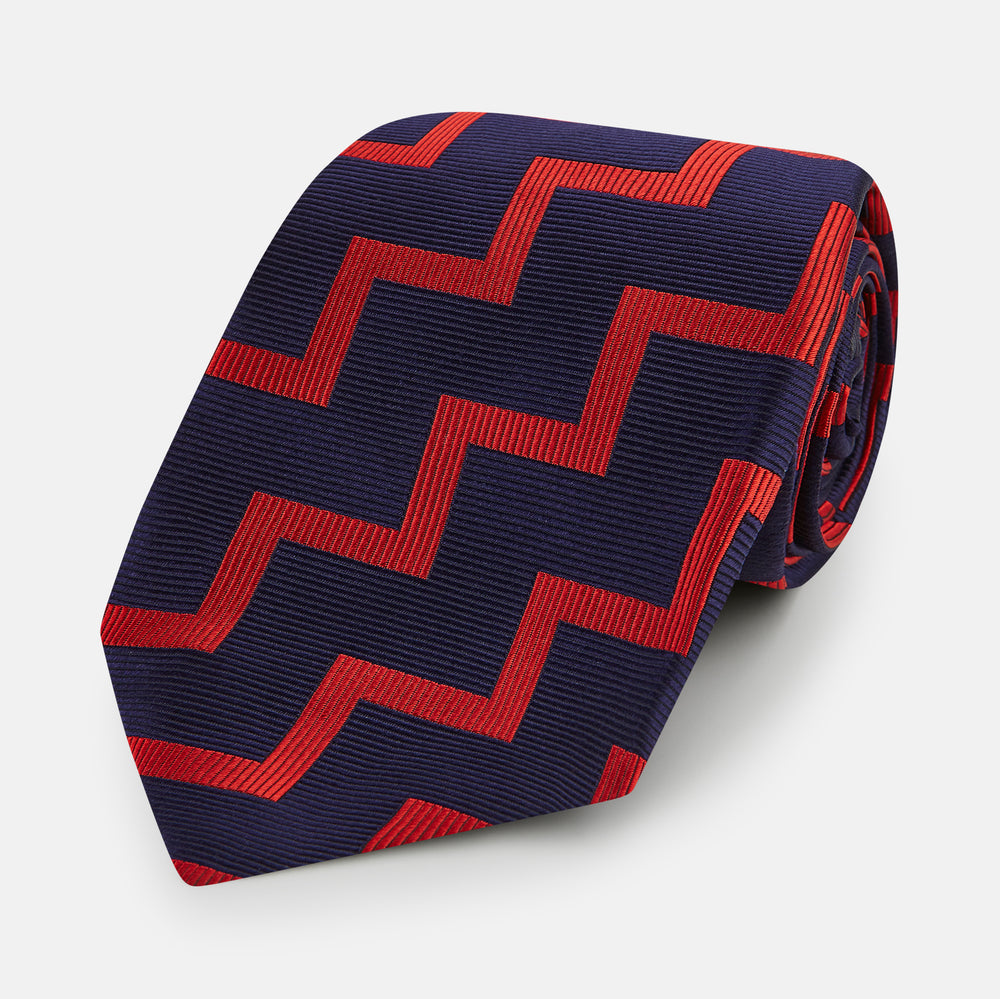 The Turnbull & Asser UK Seven-Fold Silk Red and Navy Geometric Tie is displayed rolled with its tip forward, showcasing its bold navy and red geometric pattern against a plain white background.