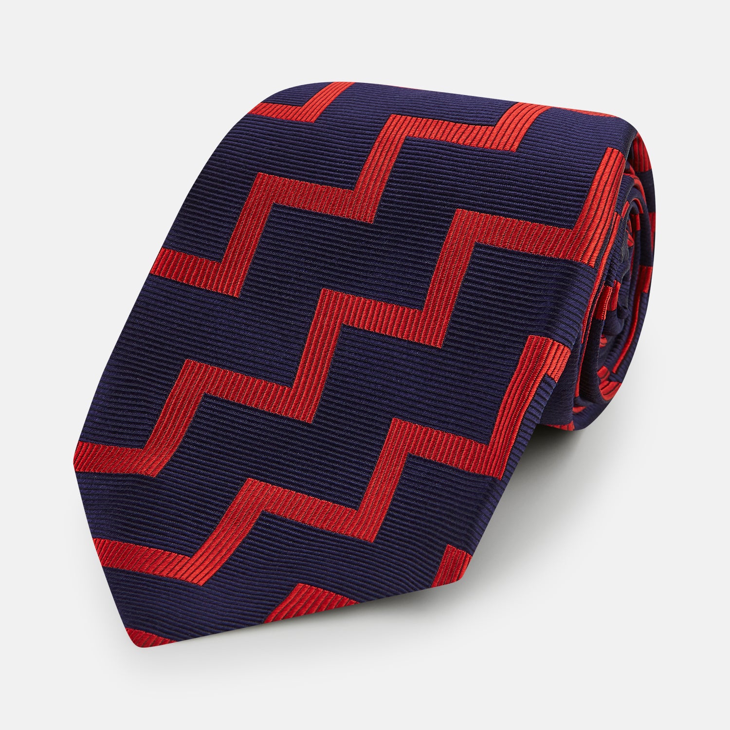 View 1 of product The Turnbull & Asser UK Seven-Fold Silk Red and Navy Geometric Tie is displayed rolled with its tip forward, showcasing its bold navy and red geometric pattern against a plain white background.