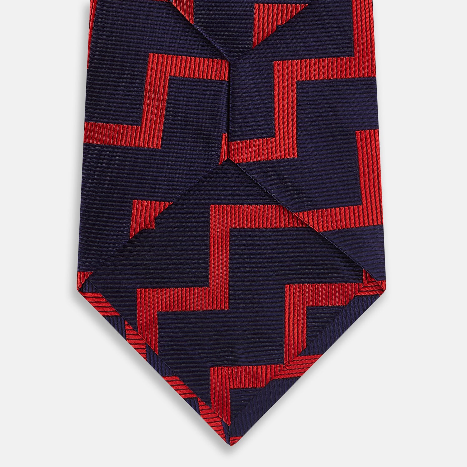 View 3 of product The Turnbull & Asser UK Seven-Fold Silk Red and Navy Geometric Tie is displayed rolled with its tip forward, showcasing its bold navy and red geometric pattern against a plain white background.