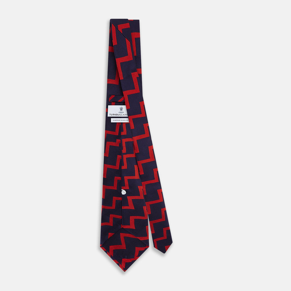 The Turnbull & Asser UK Seven-Fold Silk Red and Navy Geometric Tie is displayed rolled with its tip forward, showcasing its bold navy and red geometric pattern against a plain white background.