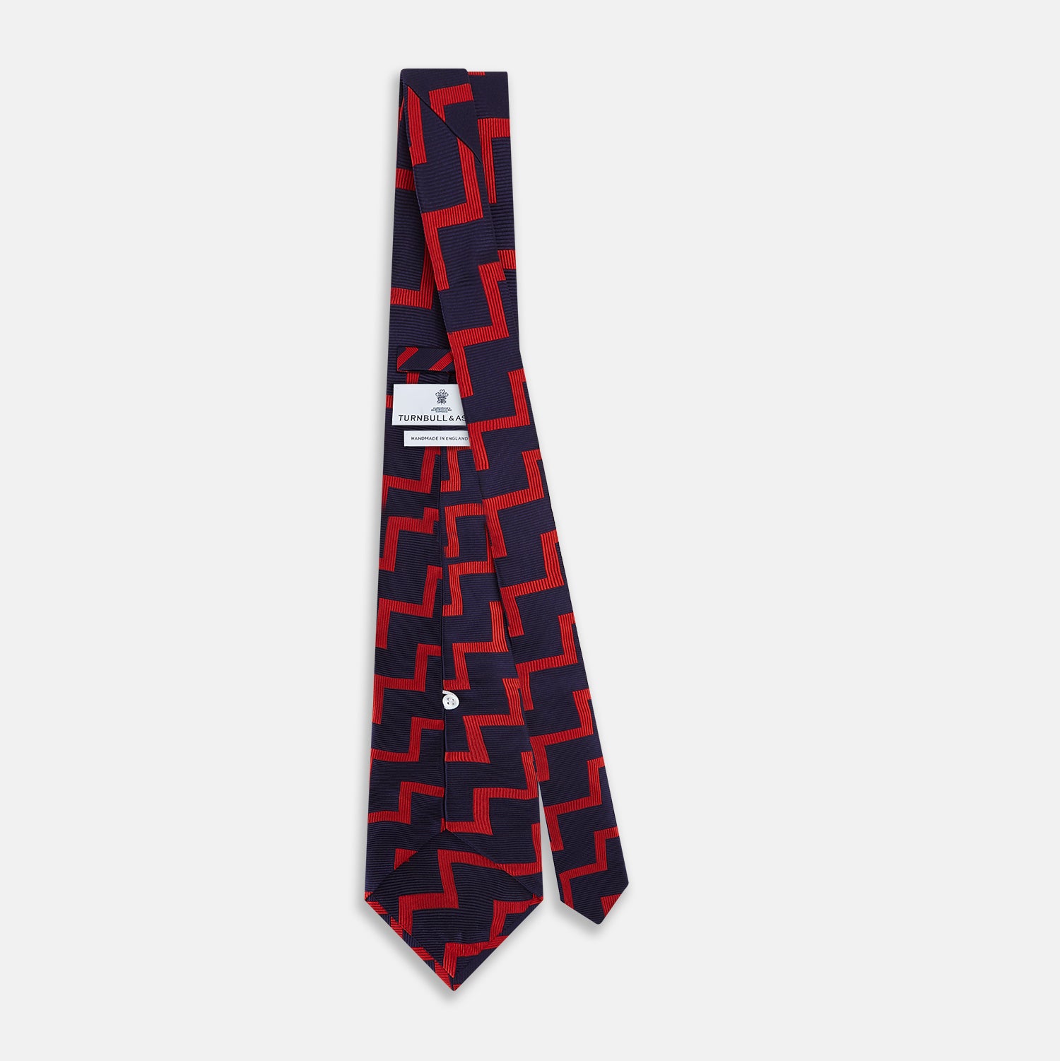 View 4 of product The Turnbull & Asser UK Seven-Fold Silk Red and Navy Geometric Tie is displayed rolled with its tip forward, showcasing its bold navy and red geometric pattern against a plain white background.