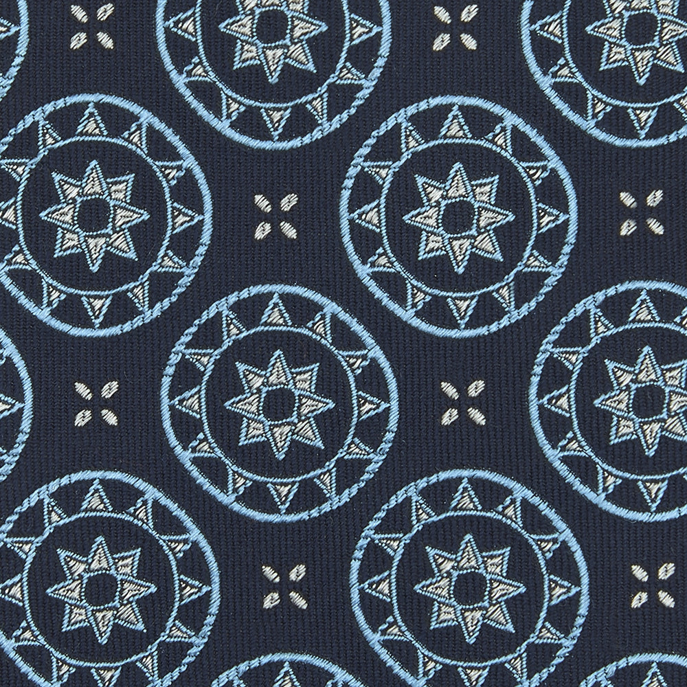 The Jacquard Silk Geometric Tie by Turnbull & Asser features a navy blue base with light blue and white star and circle motifs, shown rolled on a white background.
