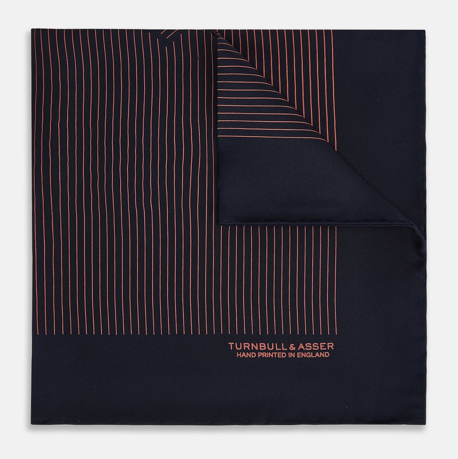 View 1 of product The Navy and Orange Stripe Silk Pocket Square by Turnbull & Asser is handmade in England from silk, featuring thin orange geometric stripes on navy and "Turnbull & Asser Hand Printed in England" printed in the corner.