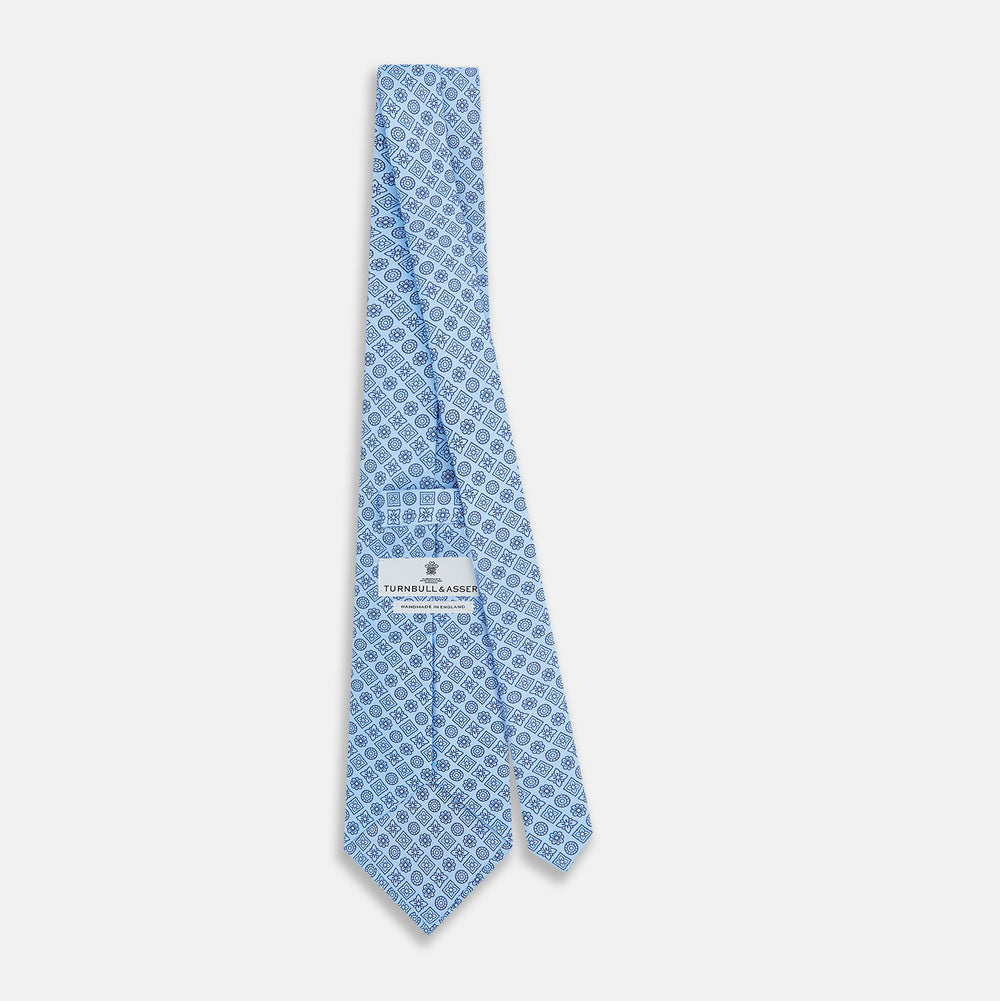 The Turnbull & Asser Blue Floral Tile Pattern Printed Silk Tie features a light blue geometric and floral design in dark blue, shown rolled up on a white background.