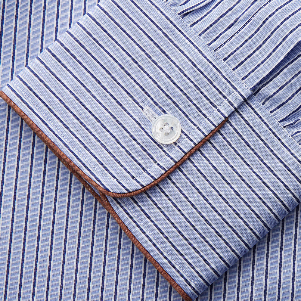 The Blue and Navy Halo Stripe Cotton Sussex Nightshirt by Turnbull & Asser features long sleeves, a collar, button-down front, chest pocket, curved hem, and brown piping for a classic luxury sleepwear style.
