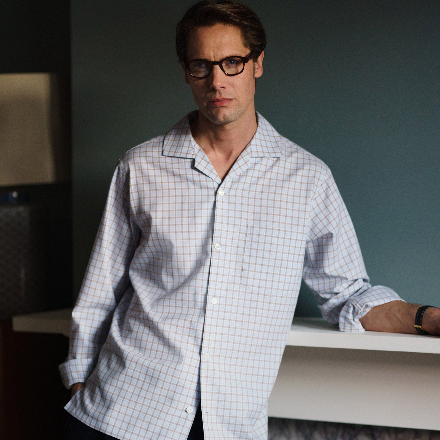 View 1 of product A man wearing glasses and a Pale Blue Multi Check Cashmerello Holiday Fit Winnington Shirt by Turnbull & Asser leans casually against a white countertop, looking at the camera with a neutral expression.