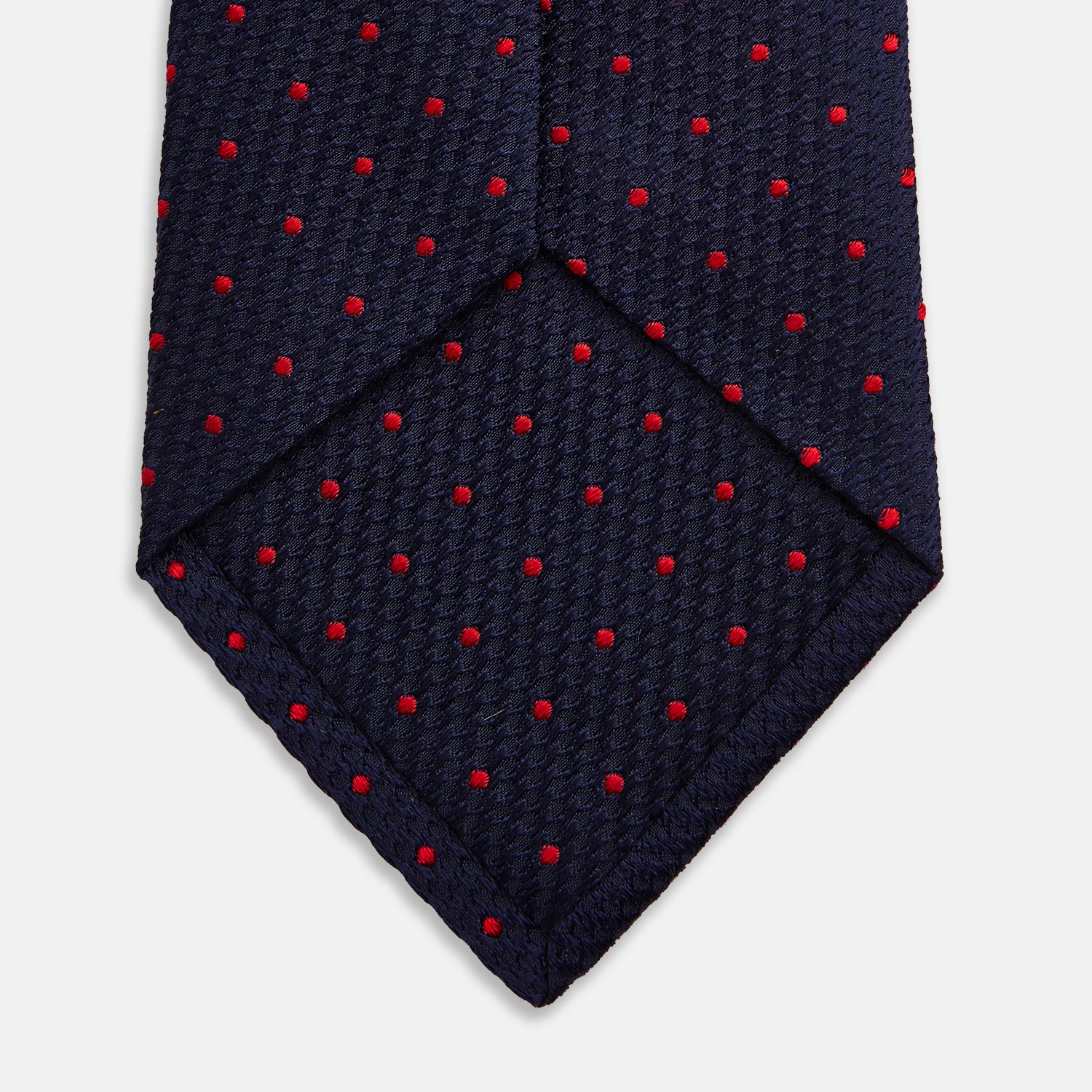 View 4 of product The Turnbull & Asser LACE ON TONE SPOT NAVY TULIP RED TIE is a classic men's accessory crafted from textured navy fabric with small red polka dots, neatly rolled with the pointed end facing forward.