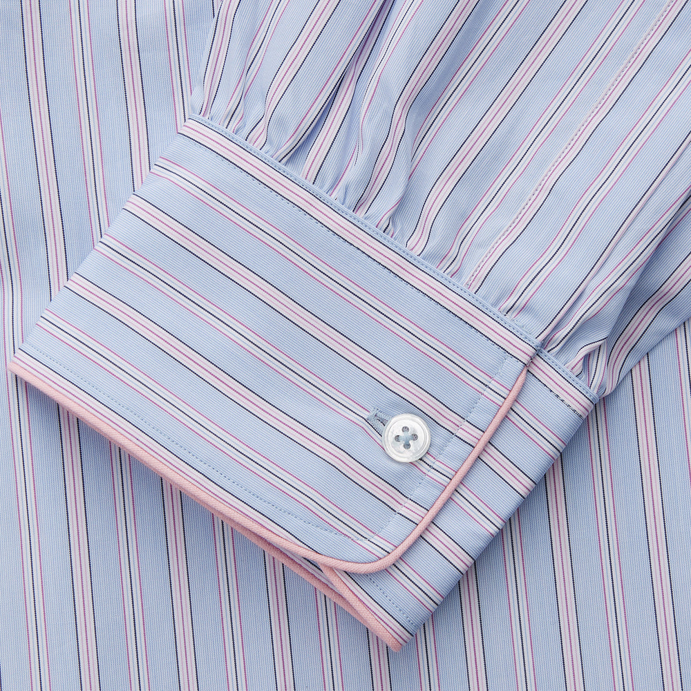 The Blue and Pink Stripe Poplin Cotton Nightshirt by Turnbull & Asser features long sleeves, a collar, front buttons, chest pocket, rounded hem, and light blue fabric with white, pink, and yellow vertical stripes.