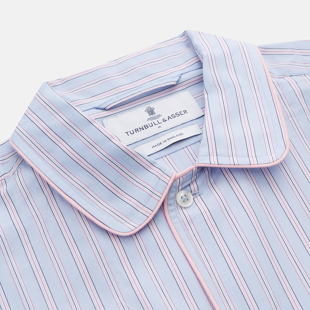 The Blue and Pink Stripe Poplin Cotton Nightshirt by Turnbull & Asser features long sleeves, a collar, front buttons, chest pocket, rounded hem, and light blue fabric with white, pink, and yellow vertical stripes.