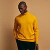 View 4 of product A man with short hair and a beard wears the Gold Aubrey Cashmere Roll Neck Jumper by Turnbull & Asser with dark pants, standing against a plain beige background and calmly looking at the camera, highlighting this stylish autumn sweater look.