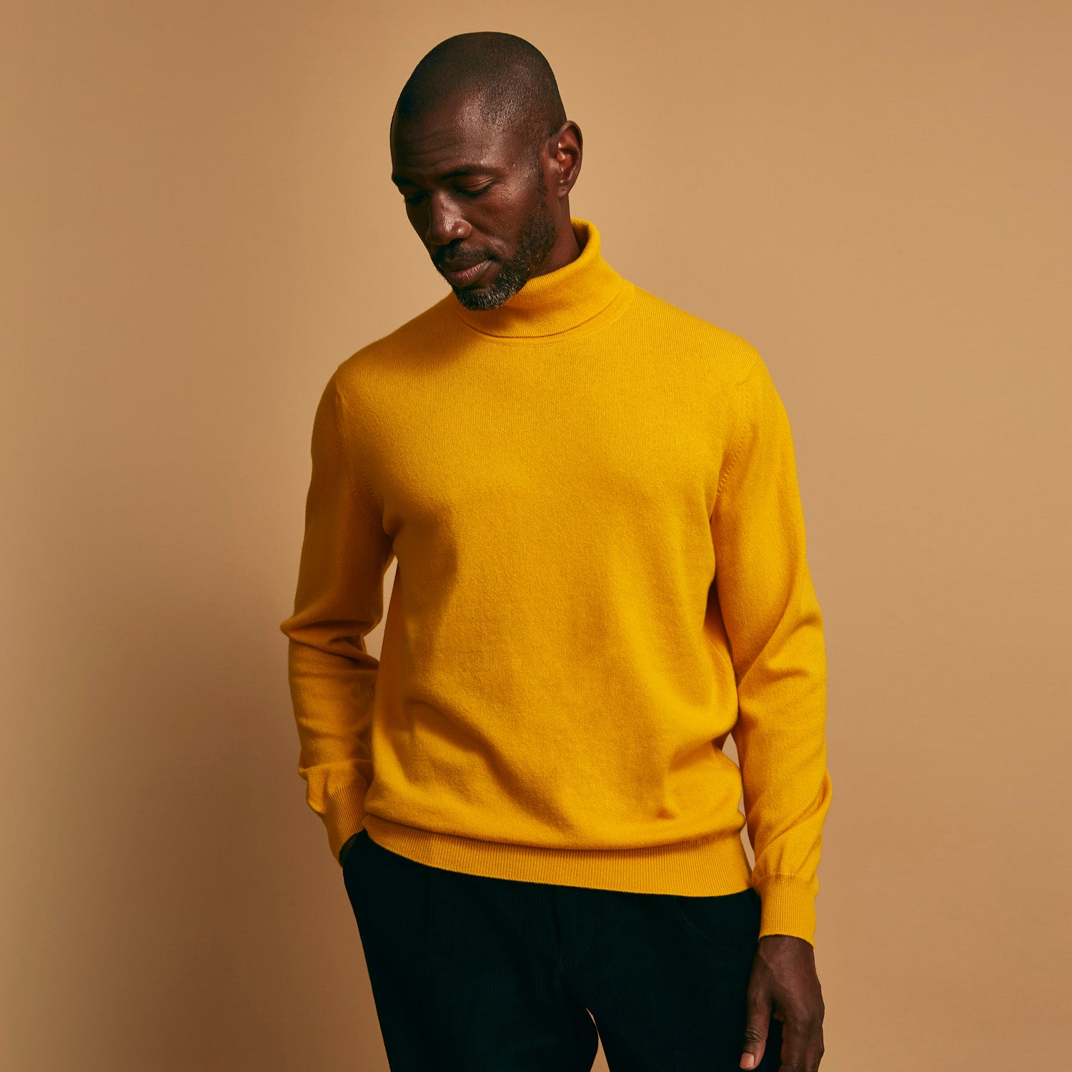 View 4 of product A man with short hair and a beard wears the Gold Aubrey Cashmere Roll Neck Jumper by Turnbull & Asser with dark pants, standing against a plain beige background and calmly looking at the camera, highlighting this stylish autumn sweater look.
