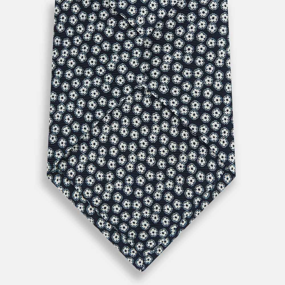 The Turnbull & Asser Navy Flower Patterned Silk Tie is shown rolled up, displaying a repeating floral design with small white and light blue circles on navy, set against a plain white background.